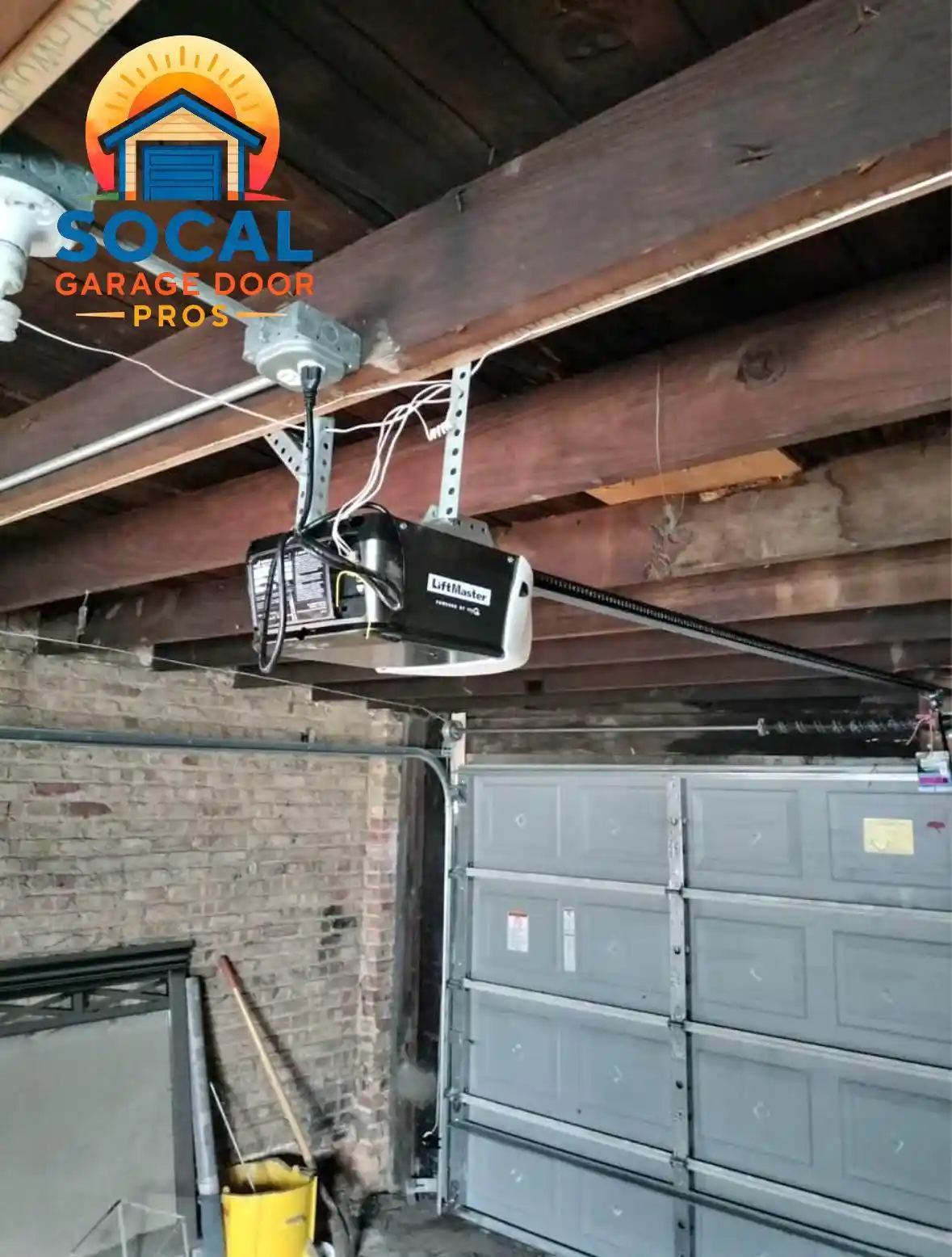 Interior of an older garage with exposed wooden ceiling beams, brick walls, and a closed gray sectional door. A ceiling-mounted garage door opener, metal rail, wiring, tracks, and torsion hardware are visible overhead, with a yellow mop bucket and stored materials along the wall.