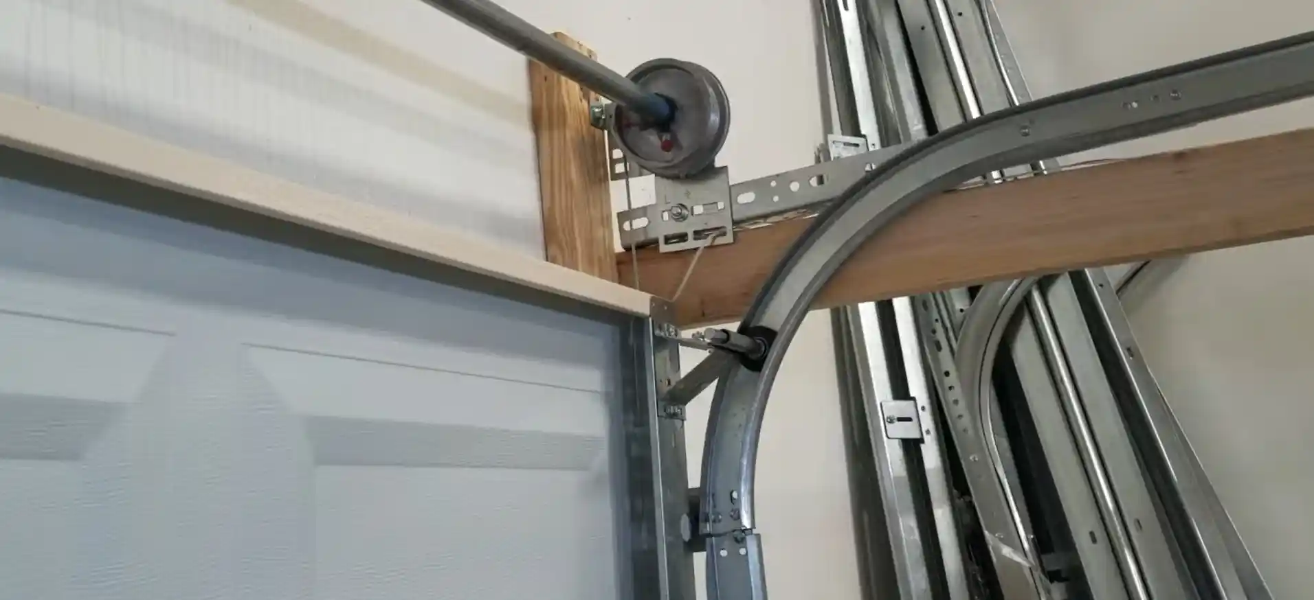 Close-up of the upper corner of a garage door showing a black roller seated in the curved metal track, with a cable drum, shaft, support brackets, and wooden framing visible nearby.