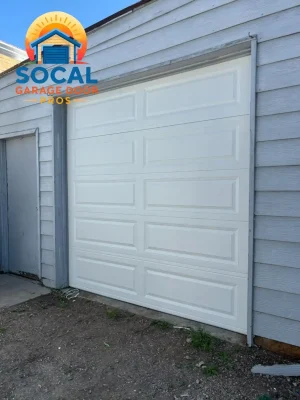 A closed white single-car garage door with raised rectangular panels is set into a building with light gray horizontal siding and unfinished trim. A plain gray side door stands to the left, and the ground in front is gravel and bare soil with small patches of grass.