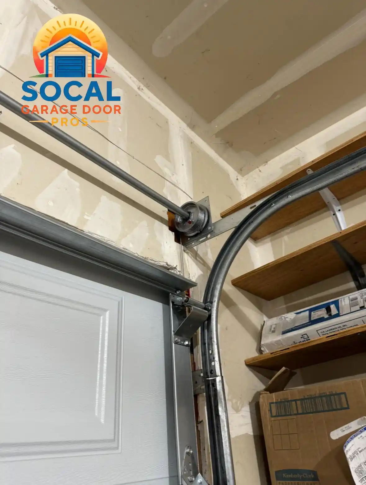 Interior corner of a garage showing the curved metal track, cable drum, shaft, brackets, and upper edge of a white sectional door. Unfinished drywall covers the walls and ceiling, while wooden shelves on the right hold boxes and packaged materials.