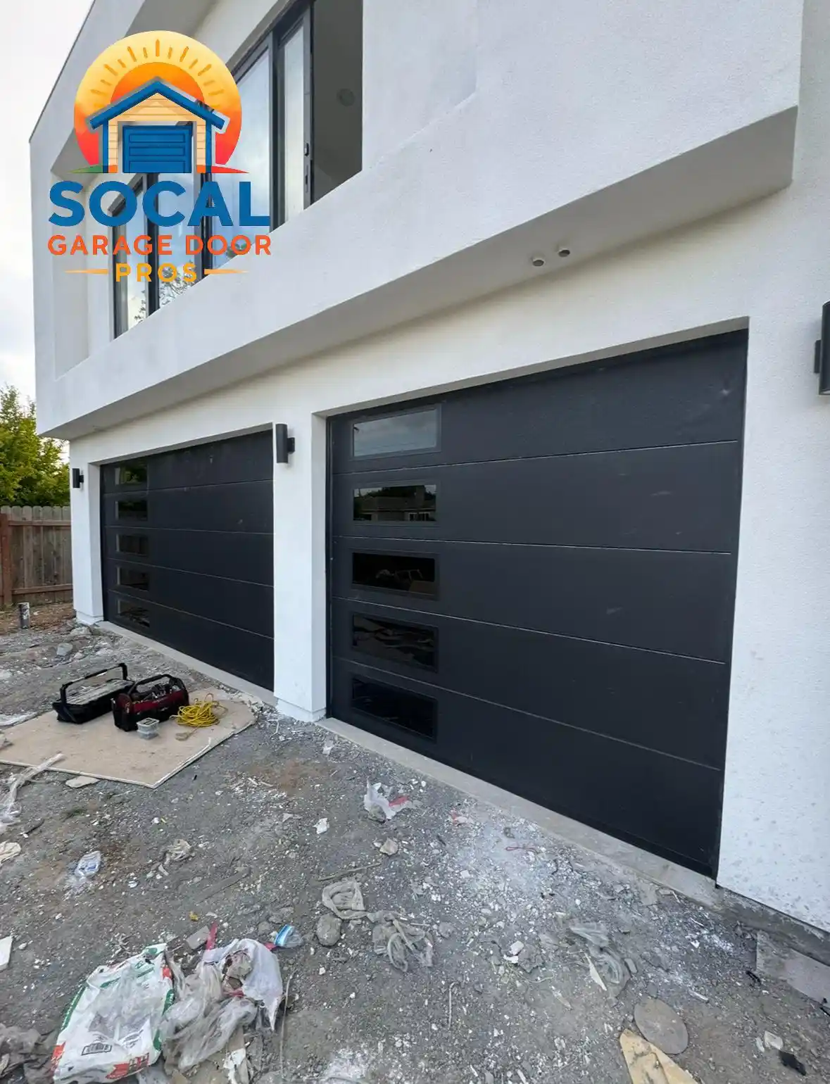 Two closed black single-car garage doors with horizontal panels and narrow rectangular windows are set into a modern white stucco building. Tools, cords, plastic sheeting, and construction debris are scattered across the unfinished ground in front.