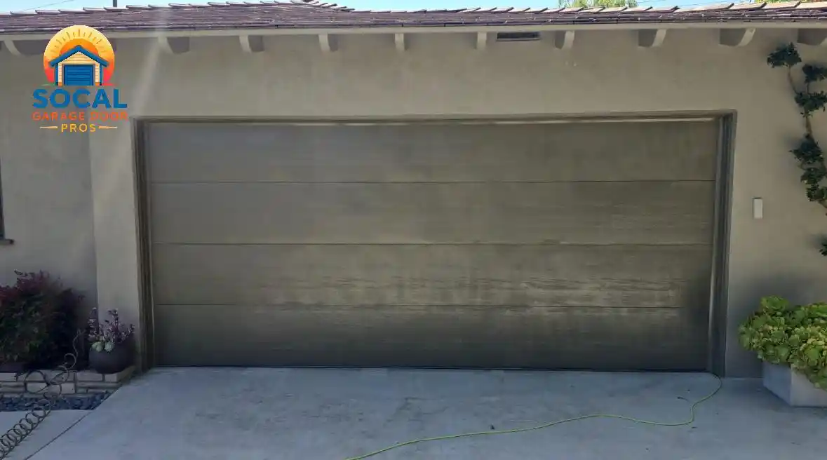 A closed dark gray double-width garage door with wide horizontal panels is set into a light stucco house beneath a tiled roof. Potted plants sit on both sides, and a green extension cord stretches across the concrete driveway.