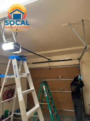 A person works near the right-side track of a closed wood-look sectional garage door. Two step ladders stand on the concrete floor beneath a ceiling-mounted opener, black rail, metal tracks, support brackets, and hanging cords.