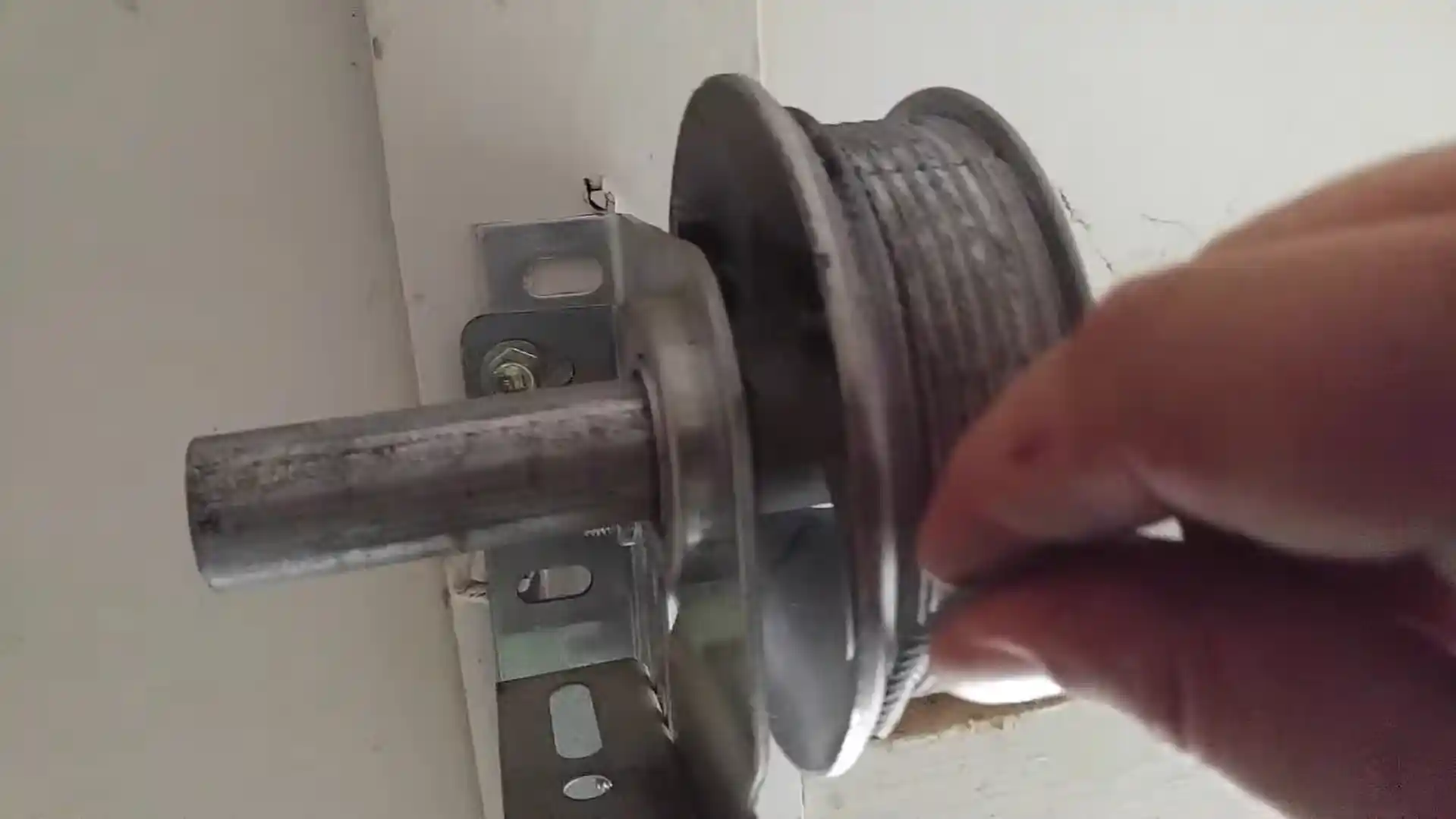 Close-up of a hand guiding a steel garage door cable onto the grooved edge of a metal cable drum mounted on a horizontal shaft and support bracket.