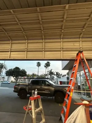 Interior of a commercial garage with a large beige sectional door partially open beneath exposed metal tracks and ceiling supports. An orange extension ladder and small step ladder stand near the doorway, while a dark pickup truck, traffic cones, palm trees, and storefronts are visible outside.