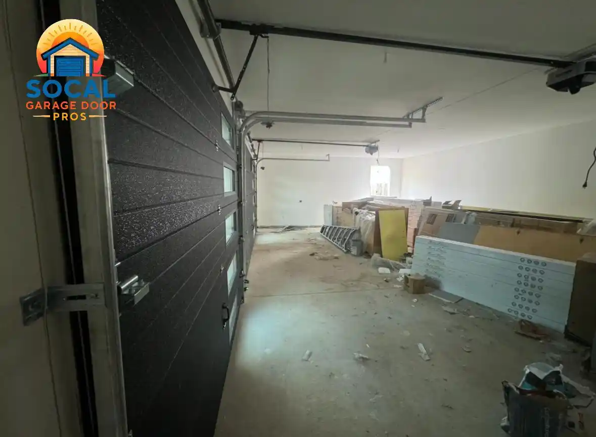Interior of an unfinished garage with a partially open dark sectional door featuring narrow rectangular windows. Metal tracks and opener hardware run along the ceiling, while ladders, boxed materials, insulation panels, and construction debris line the walls and concrete floor.