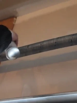 A person holds an aerosol can beneath a long garage door torsion spring mounted on a horizontal metal shaft near the ceiling.