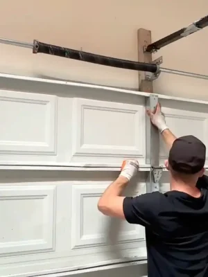 A worker wearing a black cap and white gloves aligns the upper section of a white paneled garage door. Metal reinforcement bars, hinges, side tracks, torsion hardware, and the opener rail are visible around the door.