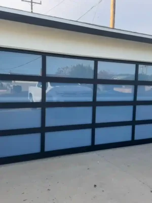 A closed modern garage door with a black grid frame and frosted glass panels is set into a white exterior wall beneath a flat roof. Reflections of parked vehicles and utility lines appear in the upper panels, and the house number “3209” is displayed above the right side.