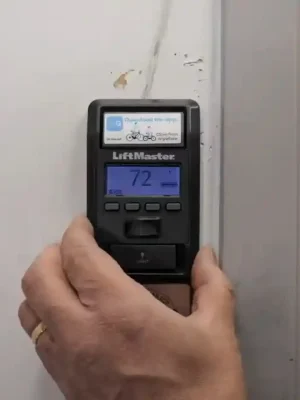 A hand holds a black LiftMaster wall-mounted garage door control beside a door frame. The blue display shows “72,” with several buttons and a small top screen visible on the device.