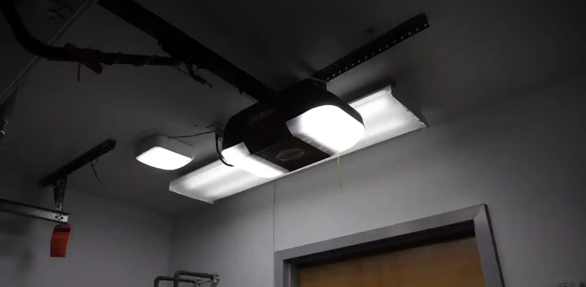 Ceiling-mounted garage door opener with two bright built-in lights, suspended from perforated metal brackets near fluorescent ceiling fixtures. A red emergency-release handle hangs from the opener rail above a doorway.