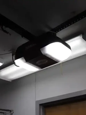 Ceiling-mounted garage door opener with two bright built-in lights, suspended from perforated metal brackets near fluorescent ceiling fixtures. A red emergency-release handle hangs from the opener rail above a doorway.