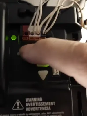 Close-up of a hand pressing a button inside the open control panel of a garage door opener. Green indicator lights, connected wires, and warning labels are visible around the controls.