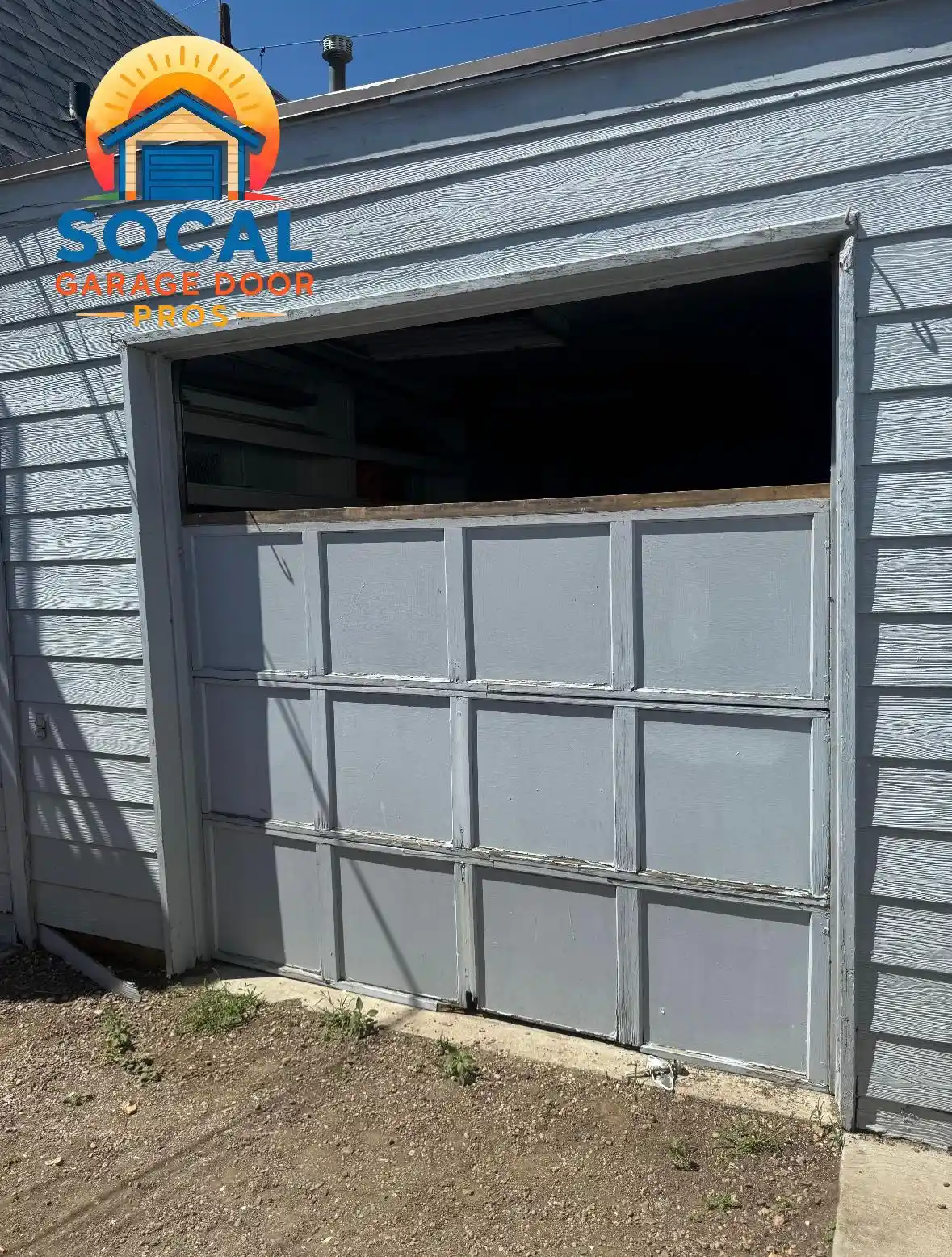 A damaged gray sectional garage door is partially open and tilted unevenly within a gray-sided building. The upper opening reveals a dark interior, while chipped paint and worn panel edges are visible above a dirt-covered ground.