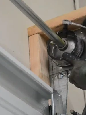 Two gloved hands use a ratchet wrench on a garage door cable drum mounted at the upper corner of the door beside the metal track and wooden frame.