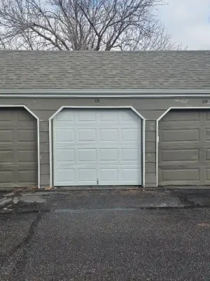 Three closed single-car garage doors are set into a gray building with white trim and a shingled roof. The center door is white, the two surrounding doors are brown, and the unit numbers “314,” “315,” and “316” appear above them.