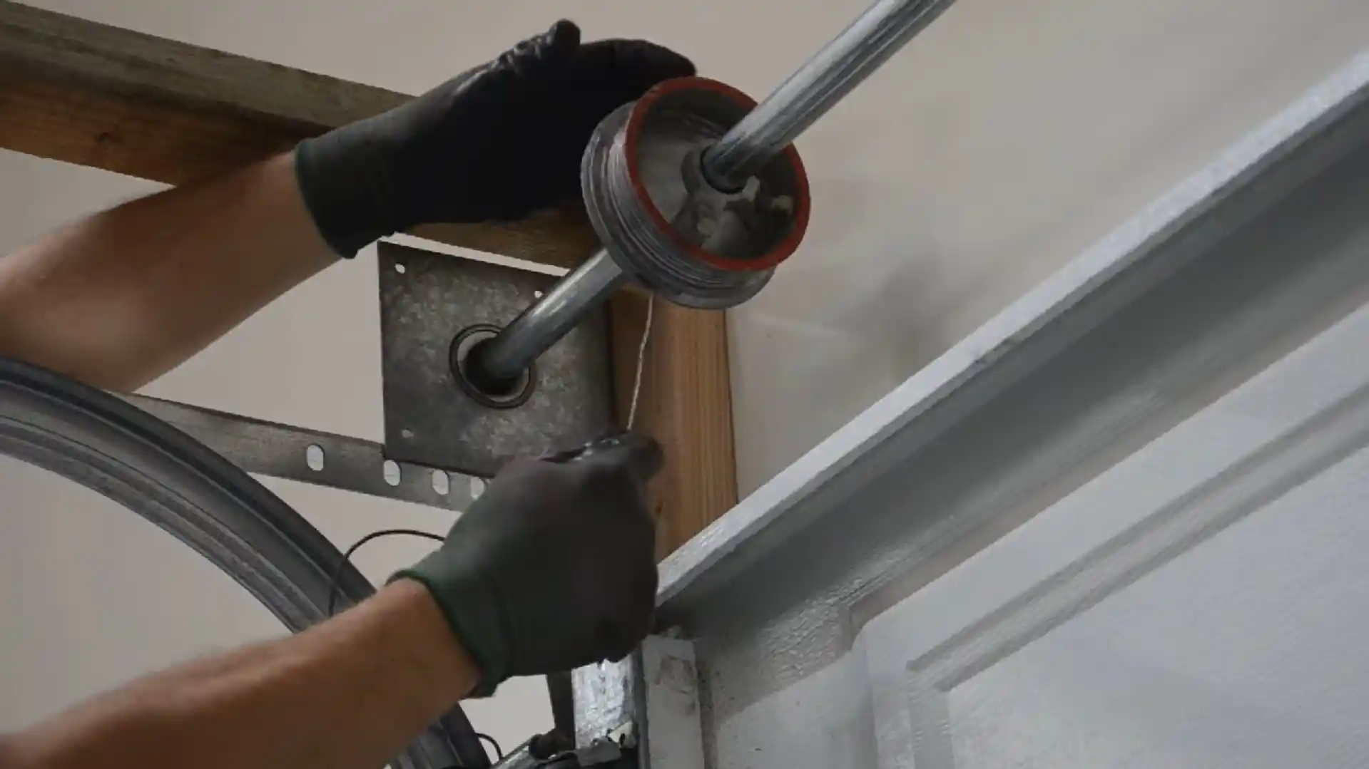 Close-up of two gloved hands guiding a thin steel cable beneath a grooved garage door cable drum mounted on a horizontal shaft near the curved track and wooden frame.