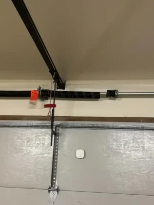 Interior close-up of the upper section of a gray garage door, showing a horizontal torsion spring and shaft mounted above it. A black opener rail, hanging emergency-release cord, metal reinforcement bracket, and small white sensor are visible.