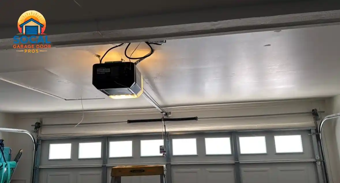 Interior view of a closed gray sectional garage door with a row of rectangular windows, metal tracks, and a horizontal torsion spring mounted above it. A ceiling-mounted garage door opener is illuminated, with wiring and a hanging release cord visible overhead.