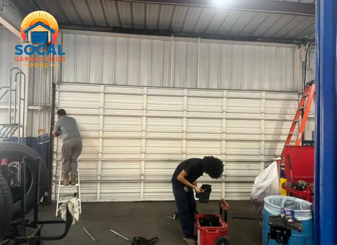 Two people work inside a metal-walled garage in front of a closed white sectional door. One stands on a small ladder near the left track, while the other bends over a red toolbox; tires, tools, ladders, bins, and equipment surround them on the concrete floor.