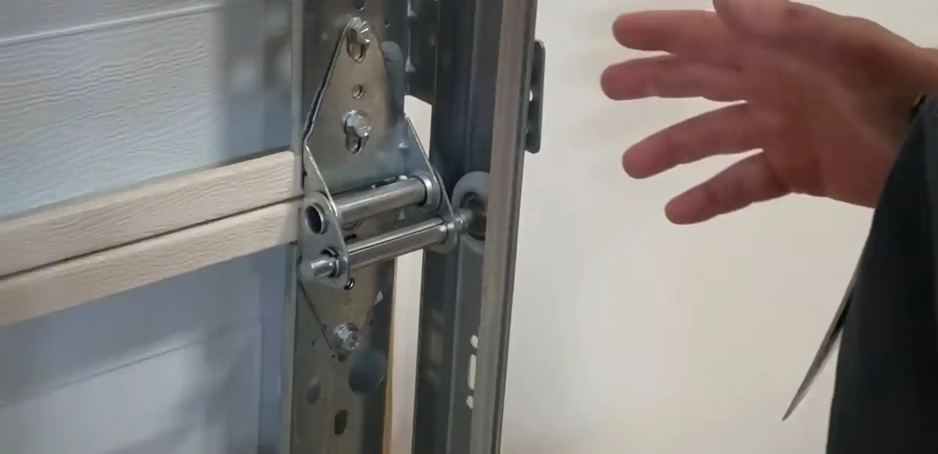 Close-up of a garage door hinge and roller assembly beside the vertical metal track, with a person’s hand positioned near the hardware.