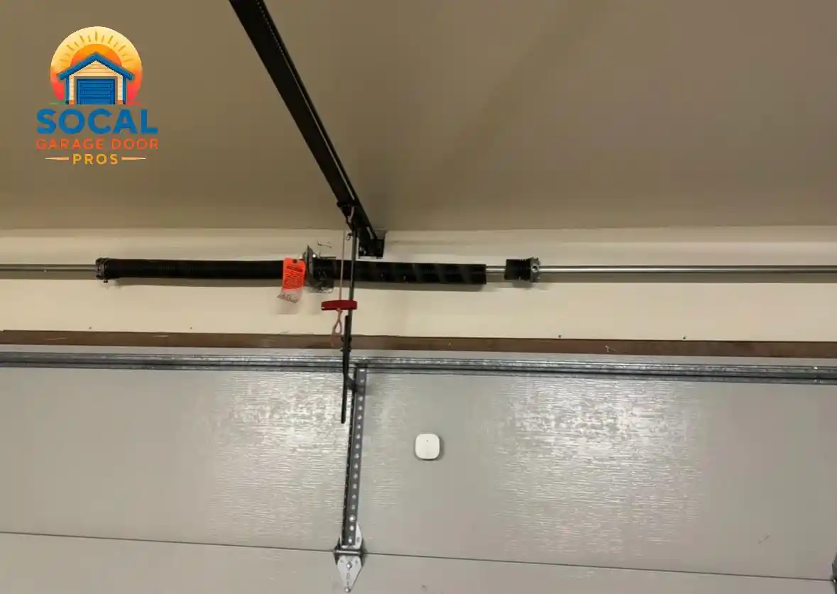 Interior close-up of the upper section of a gray garage door, showing a horizontal torsion spring and metal shaft mounted above it. A black opener rail, hanging emergency-release cord, center reinforcement bracket, and small wall-mounted sensor are visible.