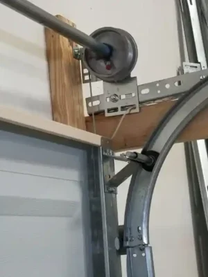 Close-up of the upper corner of a garage door showing a black roller seated in the curved metal track, with a cable drum, shaft, support brackets, and wooden framing visible nearby.