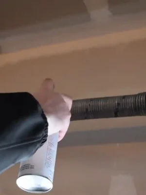 A person holds an aerosol can beneath a garage door torsion spring mounted on a horizontal metal shaft near the ceiling.