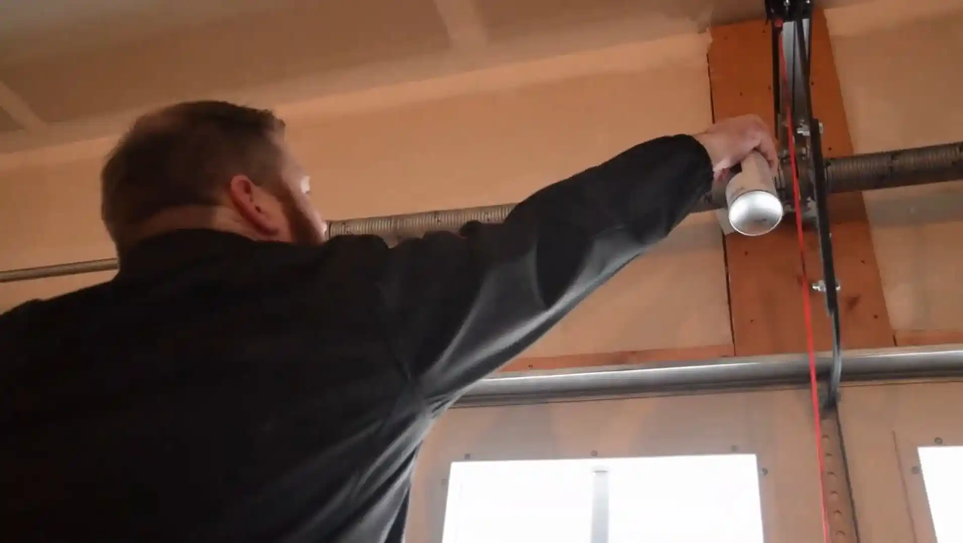 A person in a dark jacket sprays lubricant from an aerosol can onto a garage door torsion spring and metal hardware mounted above the door.