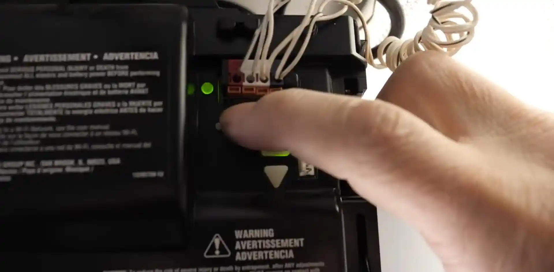 Close-up of a hand pressing a button inside the open control panel of a garage door opener. Green indicator lights, connected wires, and warning labels are visible around the controls.