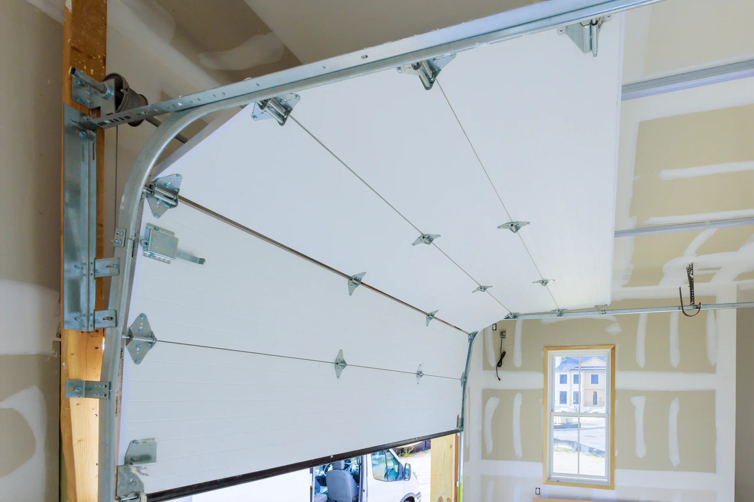 Interior view of a partially open white sectional garage door with metal hinges, cables, rollers, and curved overhead tracks. Unfinished drywall surrounds the door, and a window on the far wall overlooks neighboring houses.