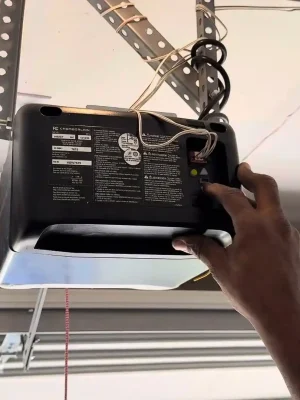 A hand presses a button on the control panel of a ceiling-mounted garage door opener. Exposed wires, indicator lights, perforated metal supports, and the garage door track are visible around the unit.