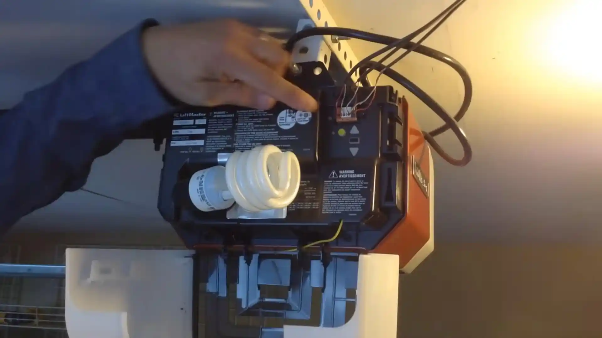 A person reaches into the exposed wiring and control panel of a ceiling-mounted garage door opener with its cover removed. A spiral light bulb, bundled cables, circuit components, and a bright illuminated bulb are visible.