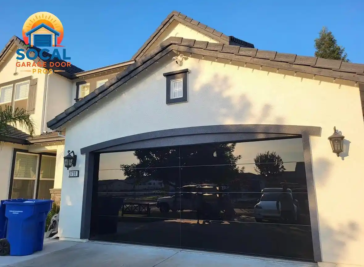 A modern black reflective double-width garage door is set into a light stucco house with dark trim and a tiled roof. The door reflects parked vehicles and nearby trees, while wall lanterns, a blue recycling bin, and the house number “7730” appear beside the driveway.