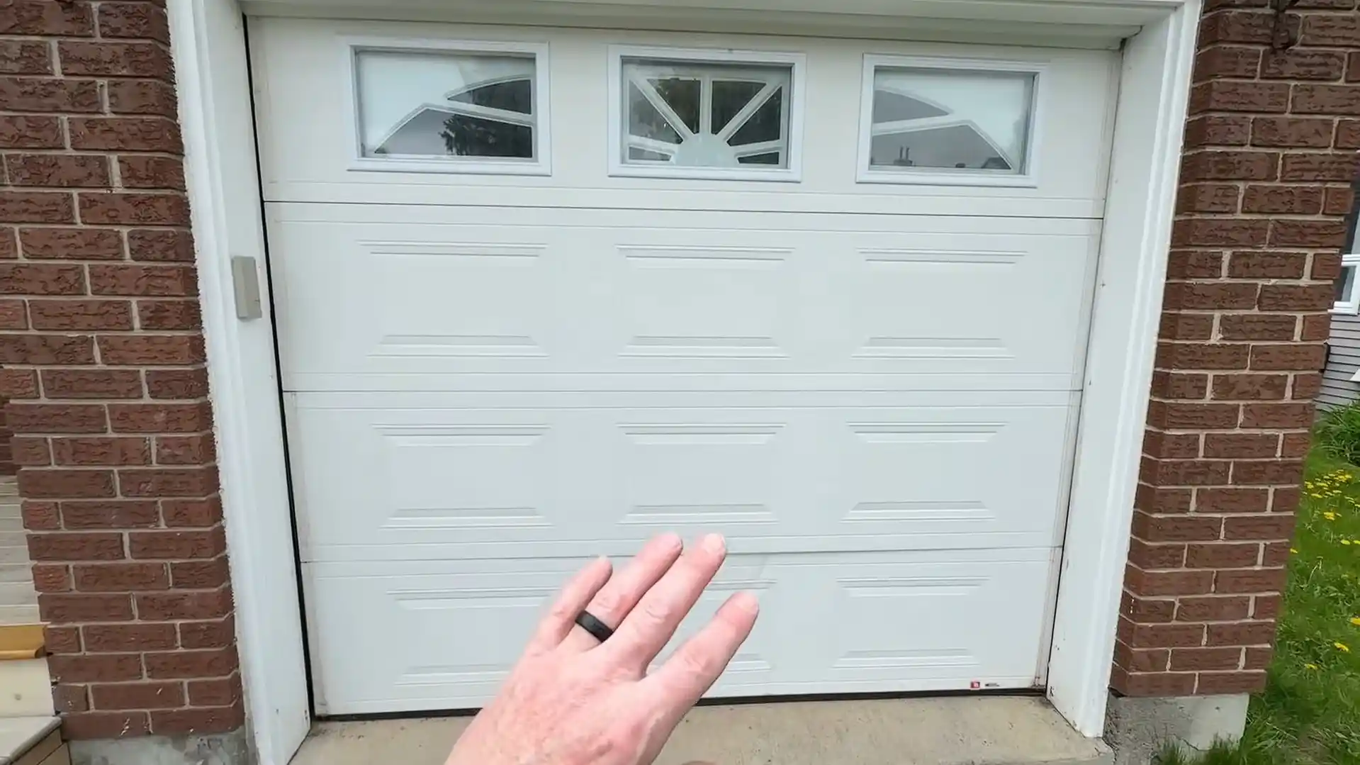 A hand gestures toward a closed white single-car garage door with raised rectangular panels and three decorative windows along the top. Red brick walls and white trim frame the door.