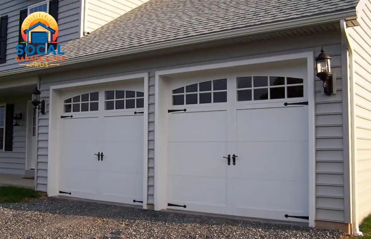 Two closed white carriage-style garage doors with arched window panels, black handles, and decorative hinges are set into a light gray-sided house beneath a shingled roof. Black lantern-style lights flank the doors above a gravel driveway.
