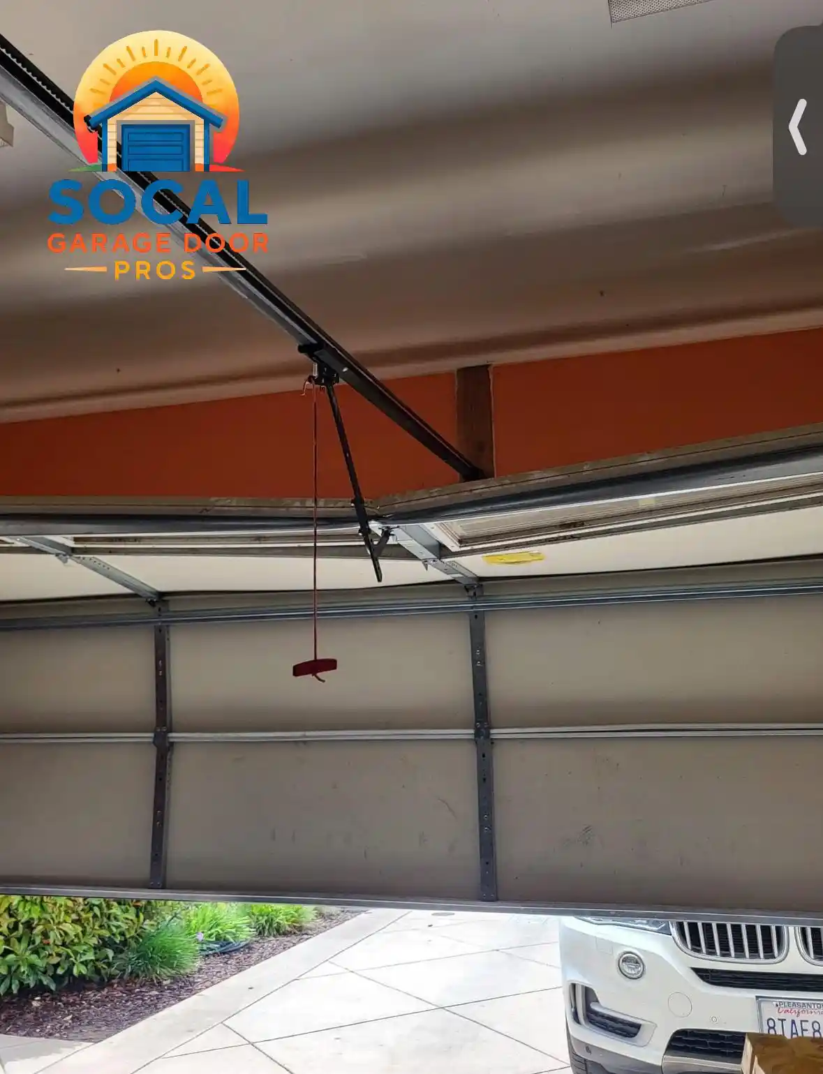 Interior view of a partially open sectional garage door with metal reinforcement rails, hinges, tracks, and a black opener arm connected overhead. A red emergency-release handle hangs near the center, and part of a white SUV is visible outside beside a landscaped driveway.