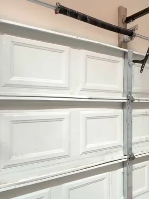 A worker wearing gloves uses a red power drill on the right-side hinge and track of a white sectional garage door. Metal reinforcement bars, rollers, curved tracks, torsion springs, and opener hardware are visible around the door.