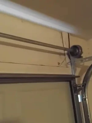 Upper corner of a garage door showing a metal cable drum on a horizontal shaft beside the curved track and support brackets. The top edge of the partially open door is visible below.
