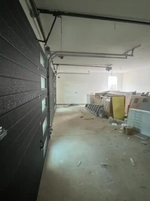 Interior of an unfinished garage with a partially open dark sectional door featuring narrow rectangular windows. Metal tracks and opener hardware run along the ceiling, while ladders, boxed materials, insulation panels, and construction debris line the walls and concrete floor.