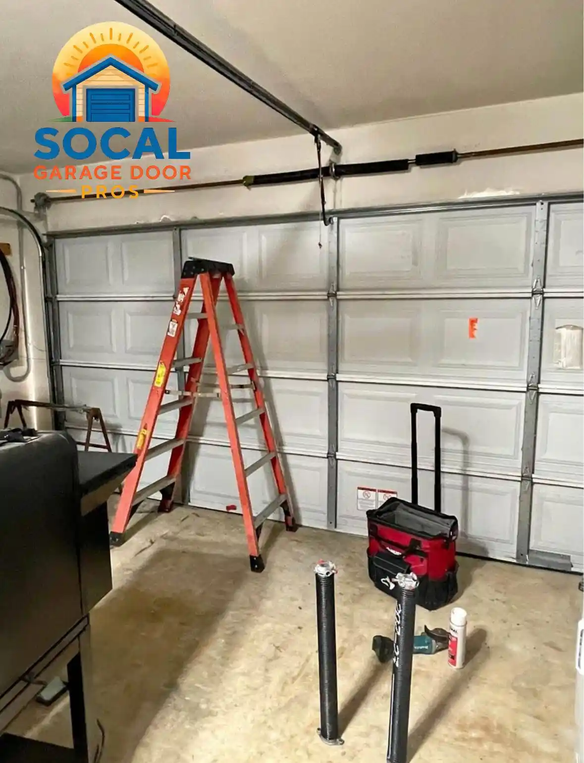 Interior of a garage with a closed white sectional door, overhead tracks, and a torsion spring assembly. A red step ladder, wheeled tool bag, power drill, spray can, and two upright black springs are arranged on the concrete floor.