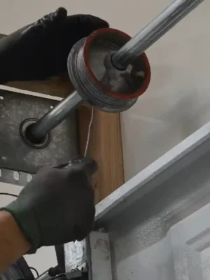Close-up of two gloved hands guiding a thin steel cable beneath a grooved garage door cable drum mounted on a horizontal shaft near the curved track and wooden frame.