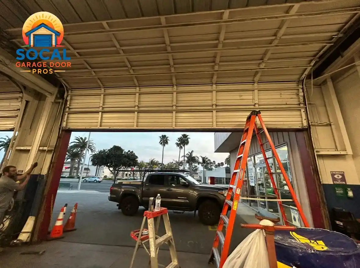Interior of a commercial garage with a large beige sectional door partially open beneath exposed metal tracks and ceiling supports. An orange extension ladder and small step ladder stand near the doorway, while a dark pickup truck, traffic cones, palm trees, and storefronts are visible outside.