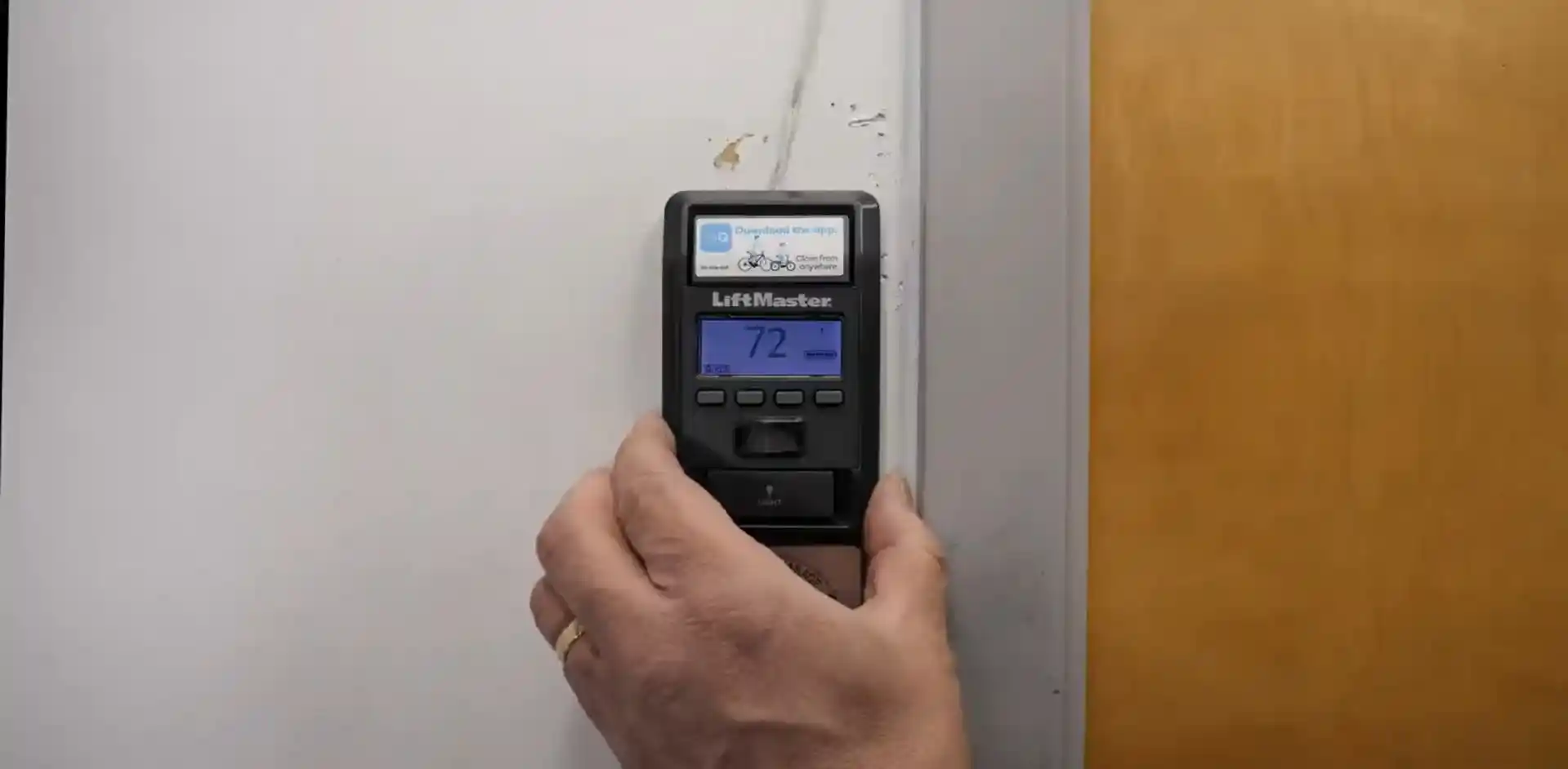 A hand holds a black LiftMaster wall-mounted garage door control beside a door frame. The blue display shows “72,” with several buttons and a small top screen visible on the device.