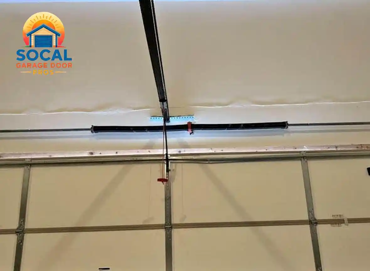 Interior view of a closed white sectional garage door with metal reinforcement rails and a black automatic opener rail extending from the ceiling to the door. A red emergency-release handle hangs from the mechanism.