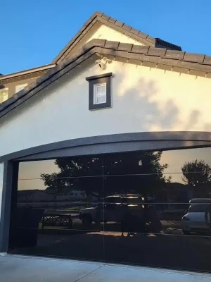 A modern black reflective double-width garage door is set into a light stucco house with dark trim and a tiled roof. The door reflects parked vehicles and nearby trees, while wall lanterns, a blue recycling bin, and the house number “7730” appear beside the driveway.