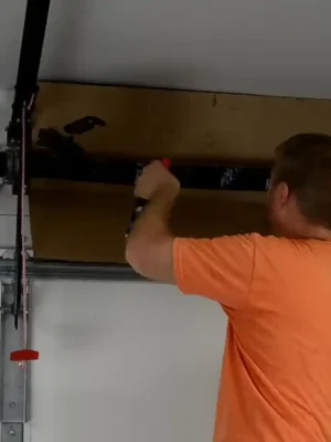 A person in an orange shirt uses a hand tool on the torsion spring assembly mounted above a white sectional garage door. A cardboard panel is positioned behind the spring and metal shaft.