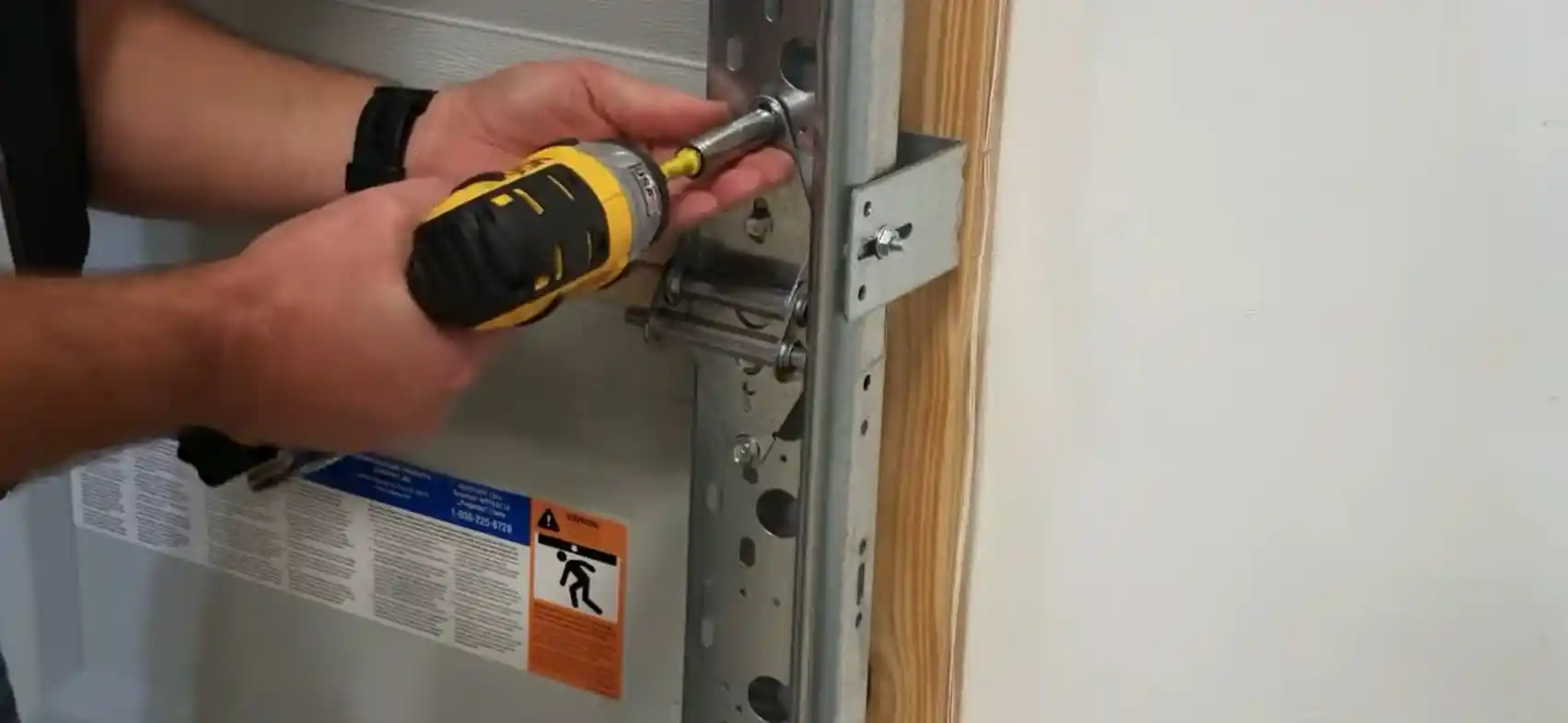 Close-up view of a technician using a yellow and black power drill to tighten a bolt on a metal bracket attached to a garage door track; the bracket is secured to a wooden frame, showing detailed hardware alignment and safety labels on the door panel.