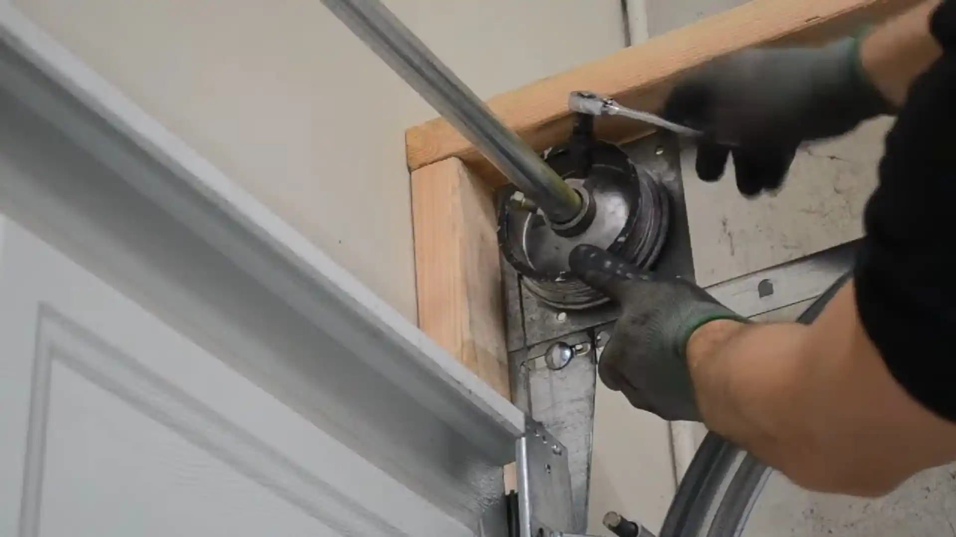 Two gloved hands use a ratchet wrench on a garage door cable drum mounted at the upper corner of the door beside the metal track and wooden frame.