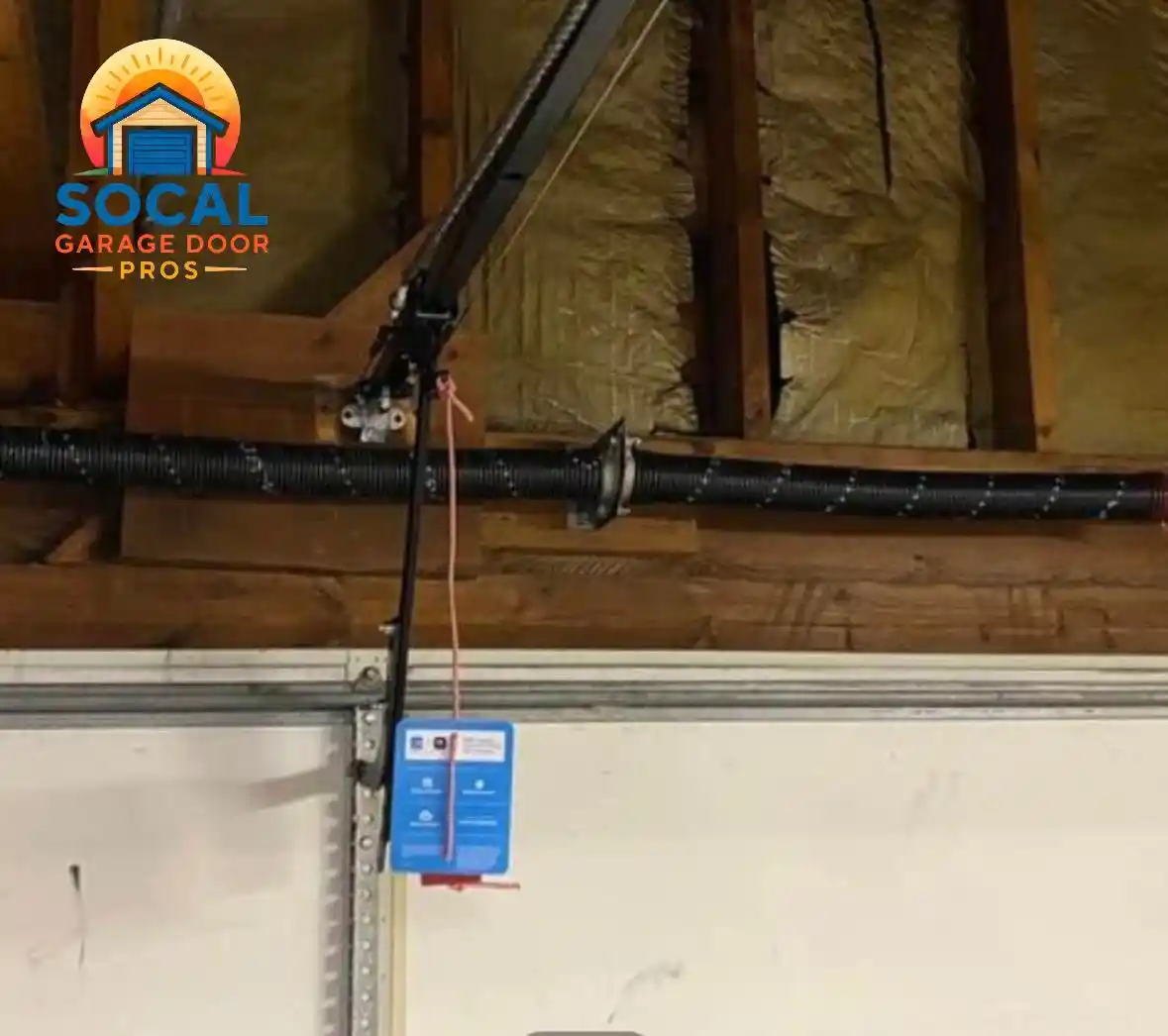Interior close-up of a garage door torsion system with two black springs mounted on a horizontal shaft above a white sectional door. An opener rail, hanging release cord, exposed wooden framing, and insulation are visible near the ceiling.